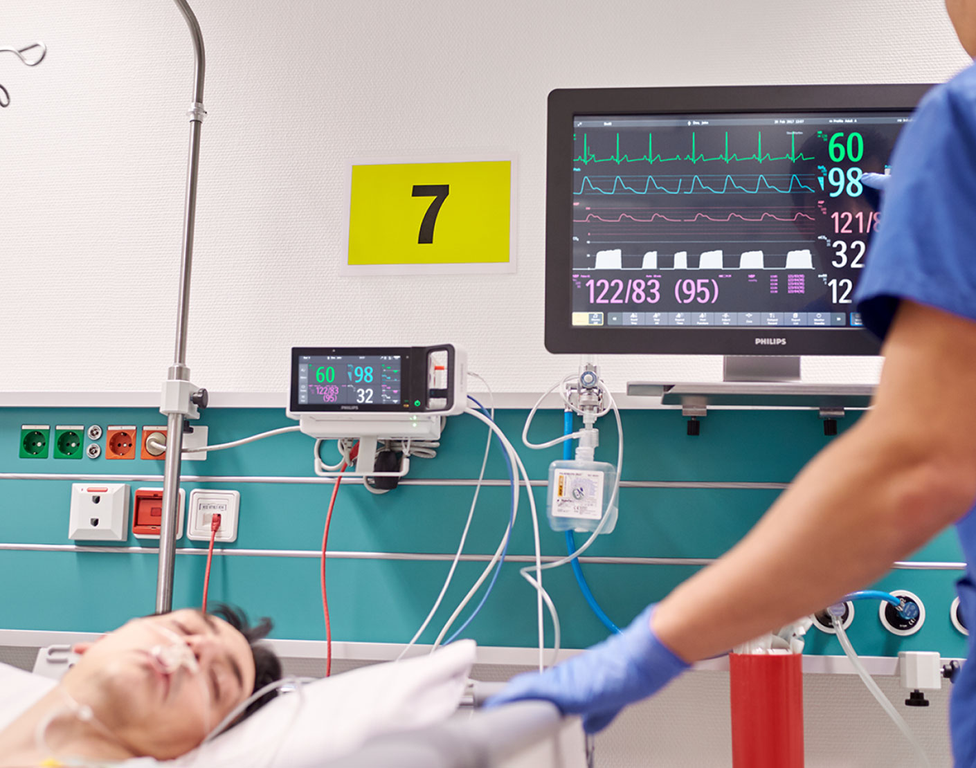 Two screens show patient status as the patient lies on bed