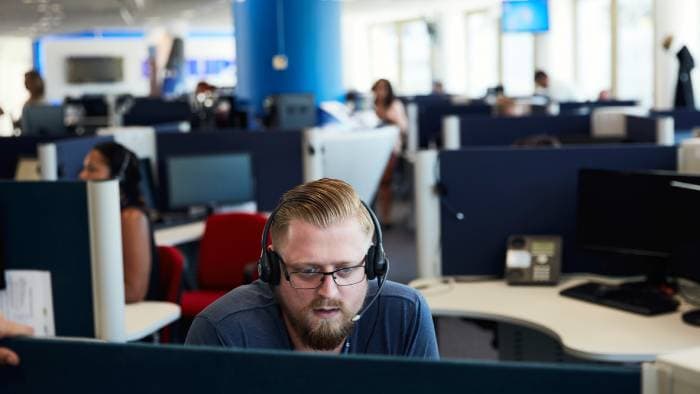 A man wearing a headset works at a desk in an busy office.
