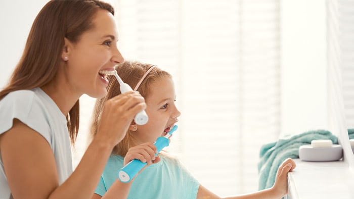 A woman and her young daughter smile while brushing their teeth with Philips electric toothbrushes.