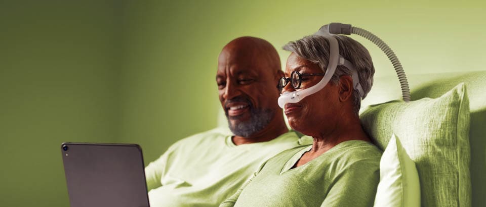 Woman in green, wearing mask sitting in bed with man watching a device