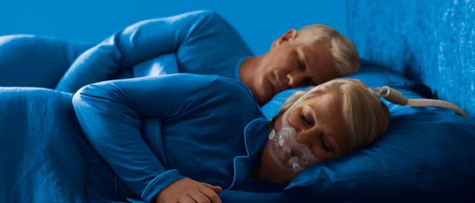Women wearing blue sleeping on her side wearing a mask and a man wearing blue sleeping on his side