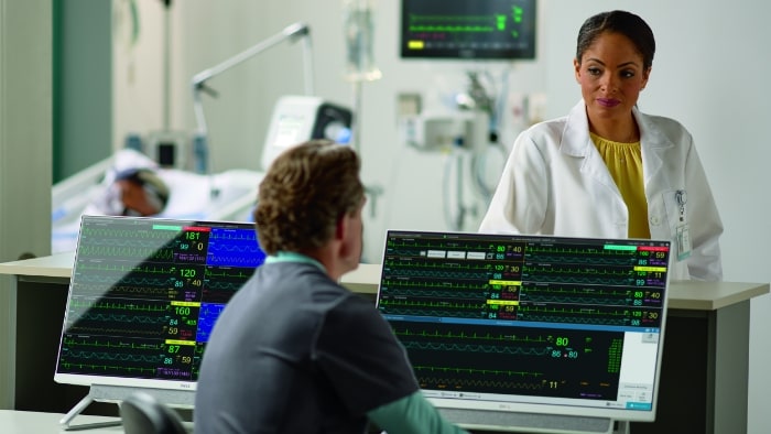 Image of healthcare providers interacting on both sides of monitor set-up, with vitals displayed.