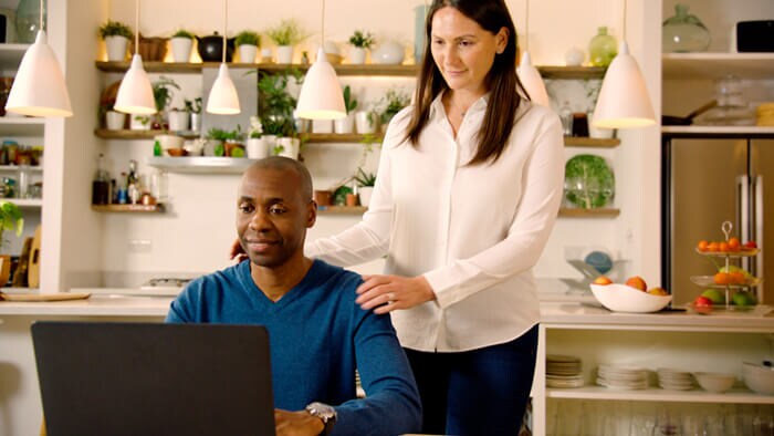 Photograph of man sat down with his wife stood up next to in the kitchen both looking at a laptop 