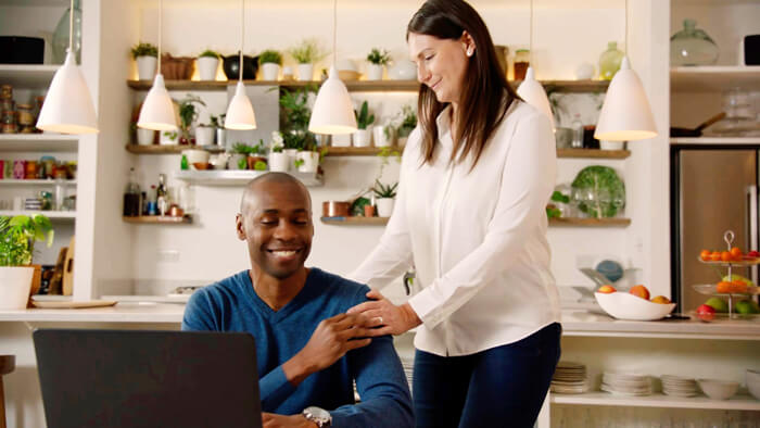 Photograph of man sat down smiling with his wife stood up next to in the kitchen smiling down at him